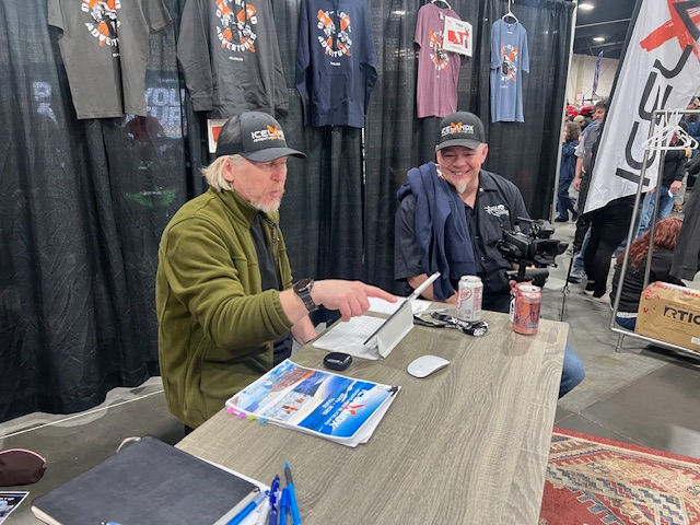 Two men in caps sit at a table with brochures and a can. Behind them, t-shirts hang against a black curtain. The mood is casual.