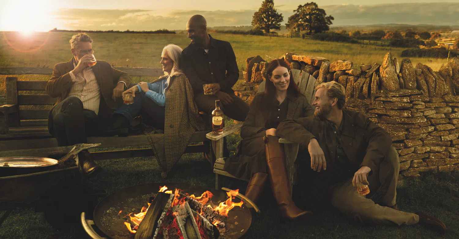 A group sat around a fire pit at sunset drinking whisky 