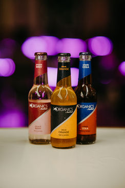 Drei Glasflaschen der Organics by Red Bull Getränke auf einem weißen Tisch, mit verschwommenem lila Licht im Hintergrund. Die Sorten sind 'Black Orange' in der Mitte, 'Purple Berry' links und 'Simply Cola' rechts. Die stilvolle Beleuchtung hebt die hochwertigen, natürlichen Erfrischungsgetränke hervor.