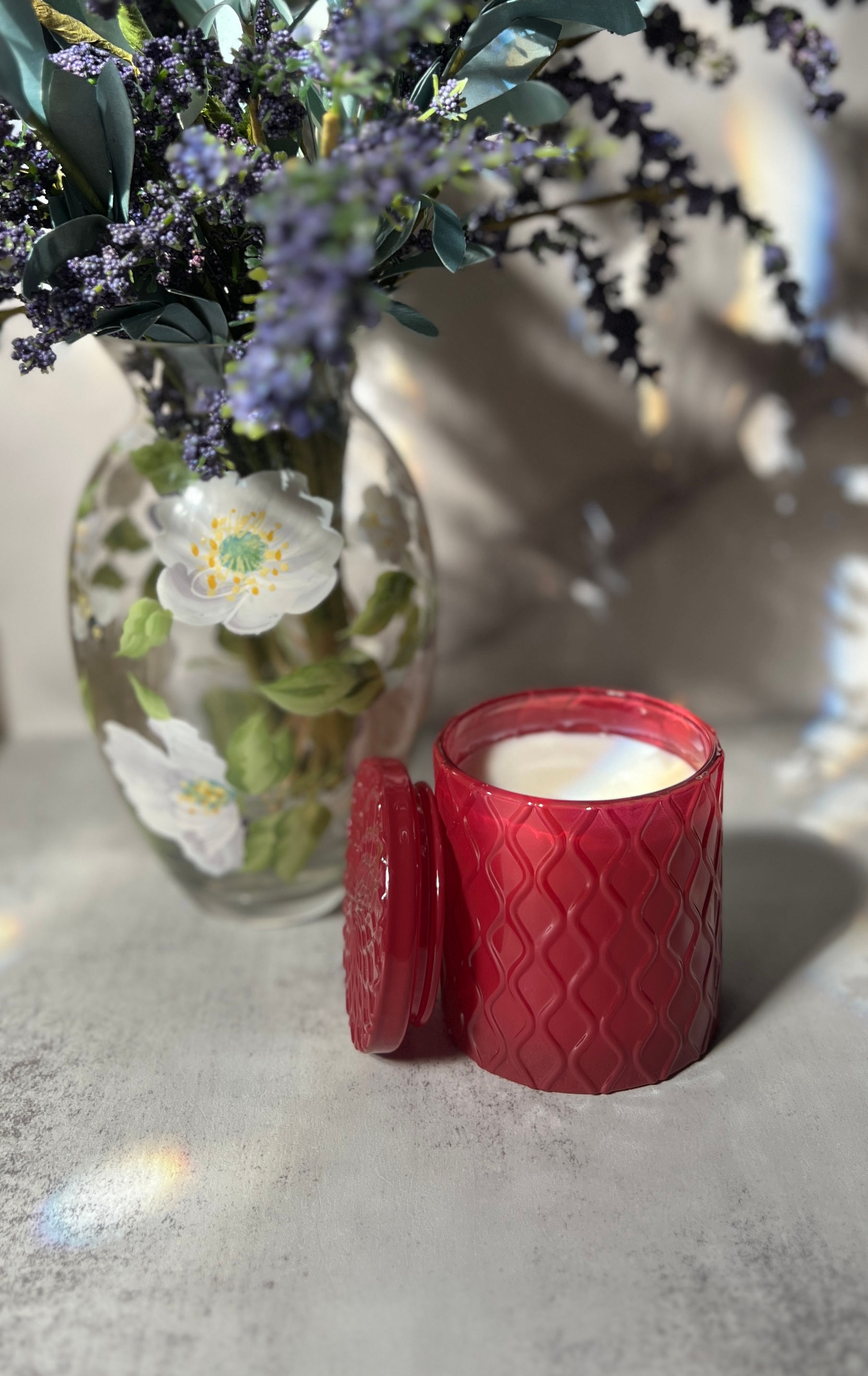 Red candle and lid beside a vase with flowers; wickless soy wax candle.