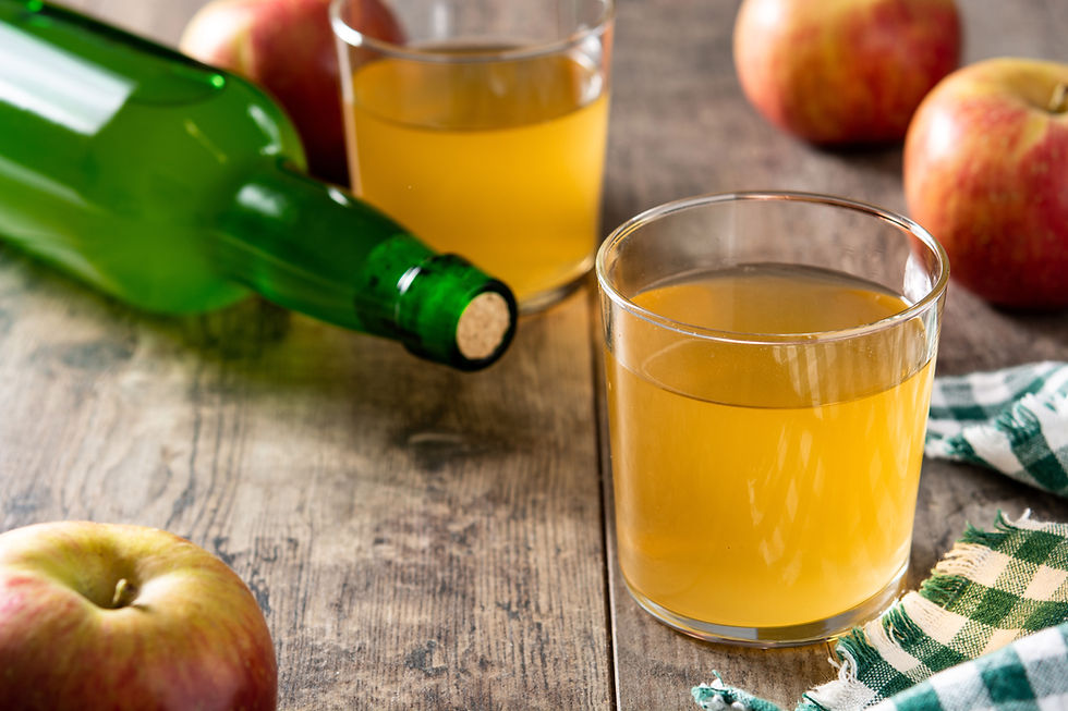 Glasses of amber cider next to a green bottle and apples on a wooden table. Green plaid cloth adds rustic charm.