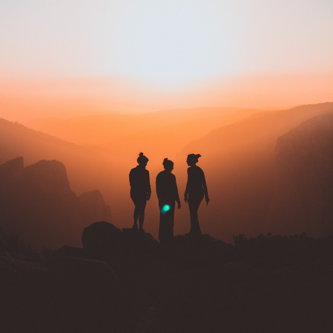 A photo of three people standing on top of a rocky outcrop with a view of mountains and a sun at dawn or dusk.