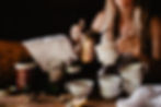 A blurry image of a woman pouring tea from a copper kettle into a mug, surrounded by many tea mugs at a table