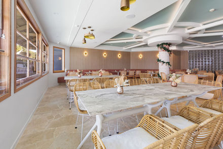 The interior of a restaurant with white walls and a timber feature wall, marble tables, rattan style chairs and a large grey tiled pillar in the middle of the room leading up to a white and blue ceiling feature