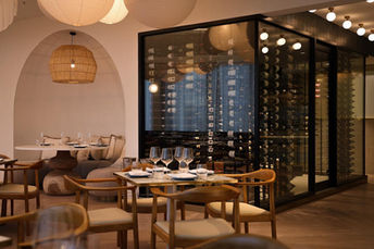 The interior of a restaurant warm lighting, timber furniture, an arched wall cutout and a glass room displaying wine