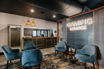 The entrance to a gaming room in a hotel with a small black marble and gold bar, upholstered blue seating, eucalyptus patterned carpet and a wall with illuminated signage reading 'Gaming Lounge' 
