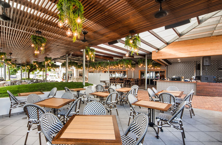 Outside seating of a hotel with timber tables and parisian chairs. The ceiling made of timber is decorated in greenery and lights.