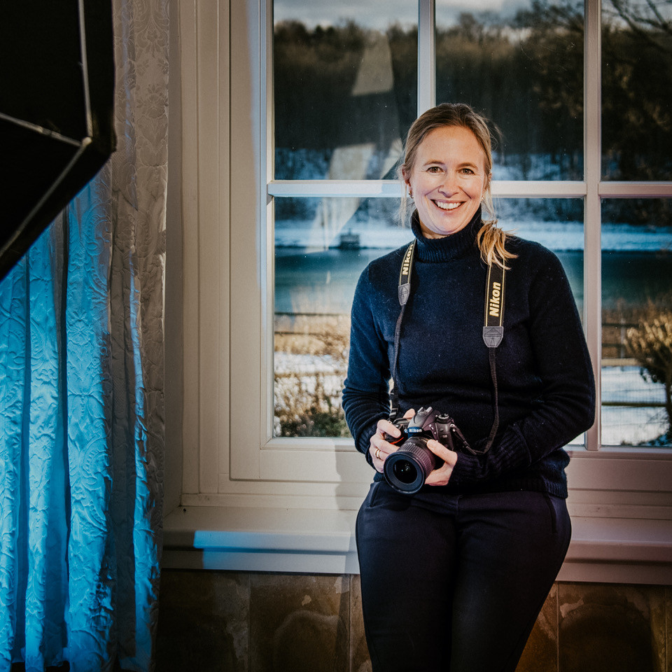 Fotografin hält lächelnd Kamera in den Händen. Sie steht vor einem Fenster
