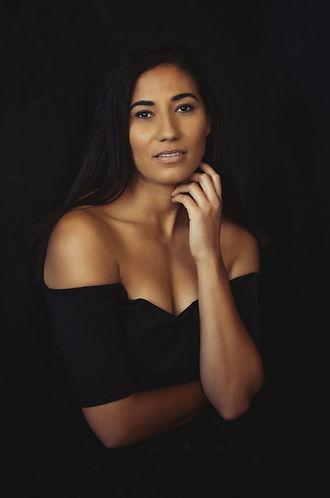 woman wearing a black dress on a black backdrop, with her hand near her face