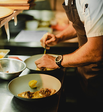Ein junger Mann mit weißer Kochjacke und brauner Schürze beugt sich über einen hellgrünen Teller und richtet darauf eine Spse an.