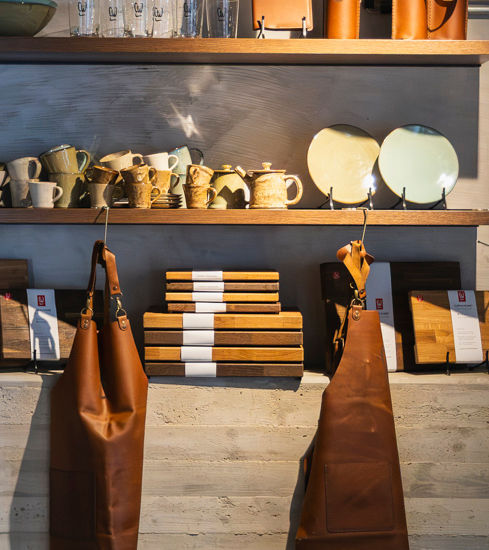 Ein kleiner Shop mit Holzregalen, die mit Schneidbretten, Mützen, Kochbüchern und Keramik bestückt sind.
