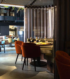 A cozy restaurant interior with a round table set for four, surrounded by orange upholstered chairs and a curved booth, softly lit by warm lighting and partially enclosed by sheer curtains.