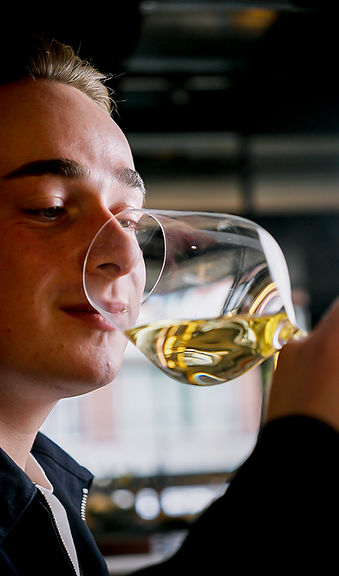 Eine Person hält ein Glas Weißwein an die Nase und scheint daran zu riechen, während im Hintergrund ein verschwommener Raum zu sehen ist.