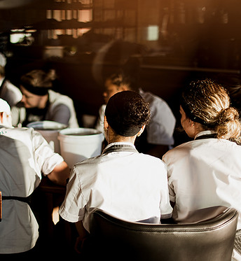 Das Bild zeigt eine Gruppe junger Frauen und Männer in Kochjacken, die um einen Tisch herum sitzen.