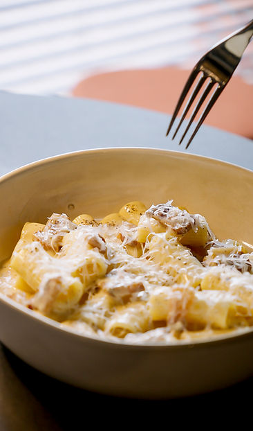 Auf einem Tisch steht eine Schüssel Rigatoni mit geriebenem Käse und Soße, darüber schwebt eine Gabel – bereit zum Essen.