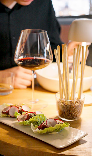 Auf einem Holztisch stehen ein Glas Rotwein, eine Schale voller Grissini und ein Teller mit Salatwraps mit Radieschenscheiben; im Hintergrund ist teilweise eine Person in schwarzer Kleidung zu sehen.