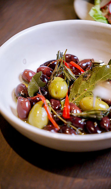 Eine weiße Schale, gefüllt mit grünen und schwarzen Oliven, garniert mit Lorbeerblättern, Kräuterzweigen und dünnen Scheiben roter Chilischoten, steht auf einem dunklen Holztisch.