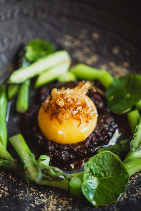 Ein Eigelb mit Knusperkrümeln auf einem runden Stück gebratenem Fleisch umringt von grünen Kräutern und Spargelstückchen.