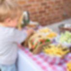 Child picks food from a table with tacos, fries, and snacks on a pink checkered cloth