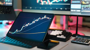 Tablet and monitors show rising stock candlestick charts on a trading desk with keyboard, calculator, and neon pink-blue glow.