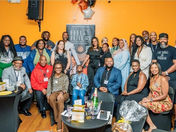 A diverse group of people posing happily in front of an orange wall with a "Feel the Future" sign. Casual attire, festive atmosphere.