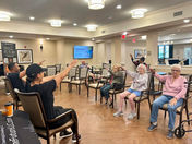 Seniors in chairs perform arm exercises in a well-lit room. A screen shows "October Birthday's." Instructors in black lead the session.