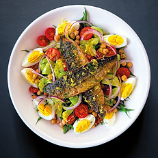 two grilled mackerel fillets covered in grated lemon zest on top of a white bowl of fresh salad of cucumber, boiled eggs, tomatoes and green beans