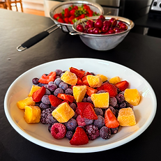 a white bowl of frozen fruit including raspberries, strawberries, blackberries, blueberries and mango, set on a kitchen worktop with two sieves in the background, one filled with cherries and the other with fresh strawberries