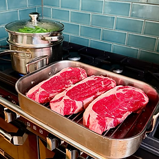 three large sirloin steaks in a steel pan waiting to be cooked next to a steamer pan full of green beans