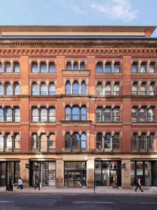 Red brick building with arched windows labeled Trinity. People walk by. Adjacent shops include a pub with signs. Clear blue sky above.