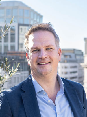 Will Kinnear smiling in a blue blazer stands outdoors with city buildings in the background under clear skies. Relaxed and professional mood.