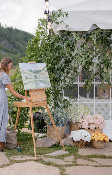 an artist sifts through paints stored in her easel. her landscape painting is on the easel and the backdrop is beautiful florals