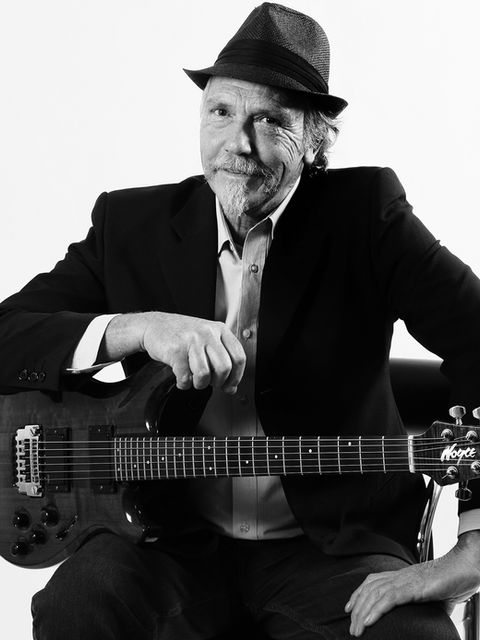 Musician Photography of an older man with a grey beared, seated on a white photo studio backdrop, holding a guitar, wearing a black suit and hat. By Musician Photographer Alise Black Studios
