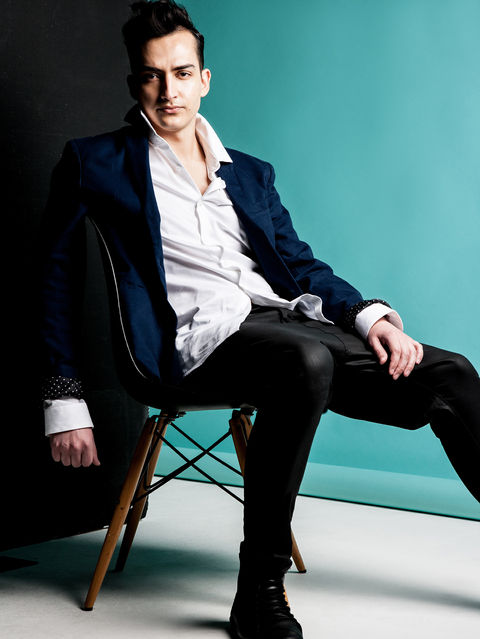 Model photoshoot of man with dark hair seated in front of teal coloured studio backdrop wearing black boots and jeans white shirt and blue jacket model portfolio by Alise Black Studios