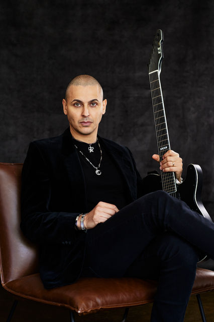 Musician Photography of a man with serious expression, wearing black jeans, top and jacket, seated on a brown chair in front of a black photo studio backdrop, holding a guitar. By Artist Photographer Alise Black