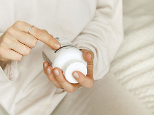 Hands in white sleeves hold a white lotion bottle, with one finger pointing at the label on a beige couch.