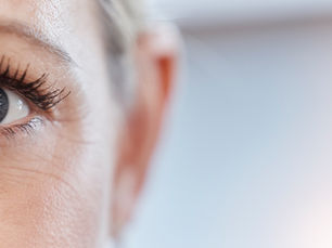 Close-up of a woman’s blue eye and face, softly lit against a blurred pale background, with a calm, attentive expression.