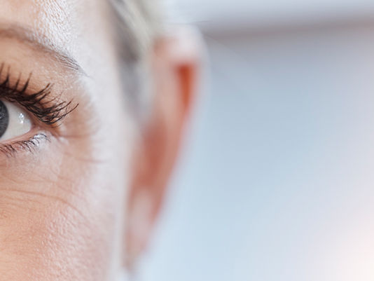 Close-up of a woman’s blue eye and face, softly lit against a blurred pale background, with a calm, attentive expression.