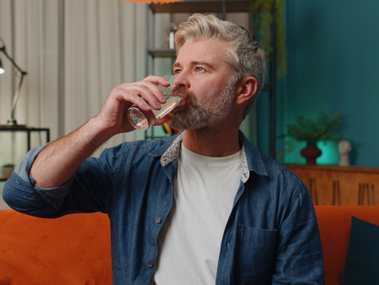 Bearded man in denim shirt drinks from a glass on an orange couch in a colorful living room with teal walls.