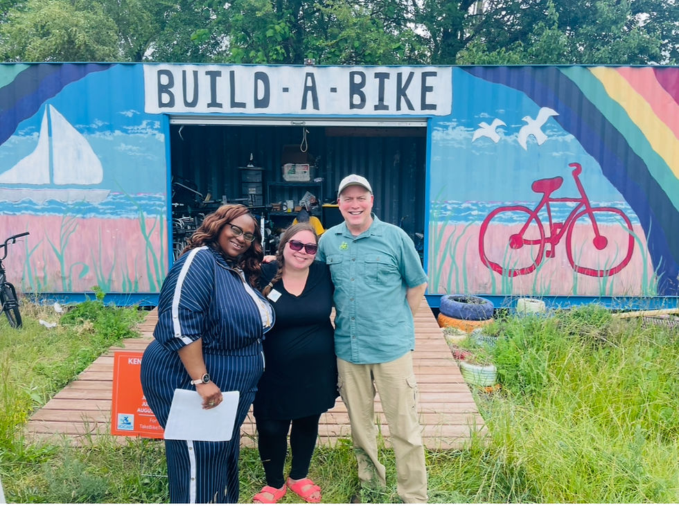     ID: NSI Cofounders, Gretchen Sipp (in a blue pin striped track suit) and Jessica Renslow (in a black sundress and leggings with pink slides), stand with NIRPC's Transportation Planning Manager / Active Transportation Planner, Mitch Barloga (in a blue button up, khakis and a ballcap before The Ken Parr Build a Bike Popup Shop. 