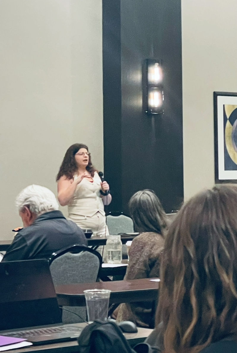 ID: Literary Agent, Rachel Estep, dressed in a white pantsuit, holds a mic and describes what is on her manuscript Wishlist. 