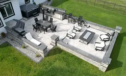 Full rectangular patio with walls and a fire pit. There is a sitting area surrounding the fire pit and then another one with a table near the full service outdoor kitchen. There are paver stairs going up to the door and a hot tub opposite the stairs.