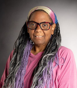 Black woman with a multicolored headband, purple/grey braids, wearing a pink shirt. She is wearing multicolored purple glasses, sitting with a grey background.