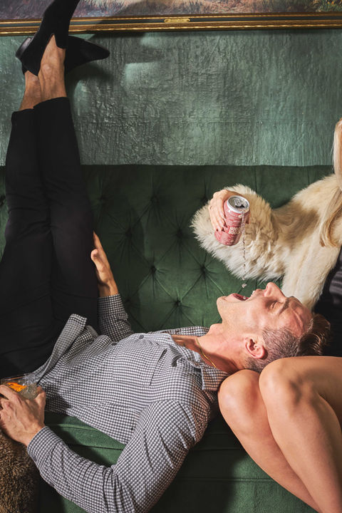 Man laughs on couch with a drink being poured into his mouth
