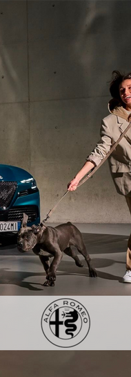 Donna che porta a spasso un cucciolo di Cane Corso, entrambi vicino a un'auto blu Alfa Romeo. Pubblicità con cane, Animal Spot Milano.