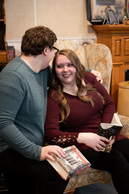 man whispers something to fiance, they're sitting on an old, high back chair holding books