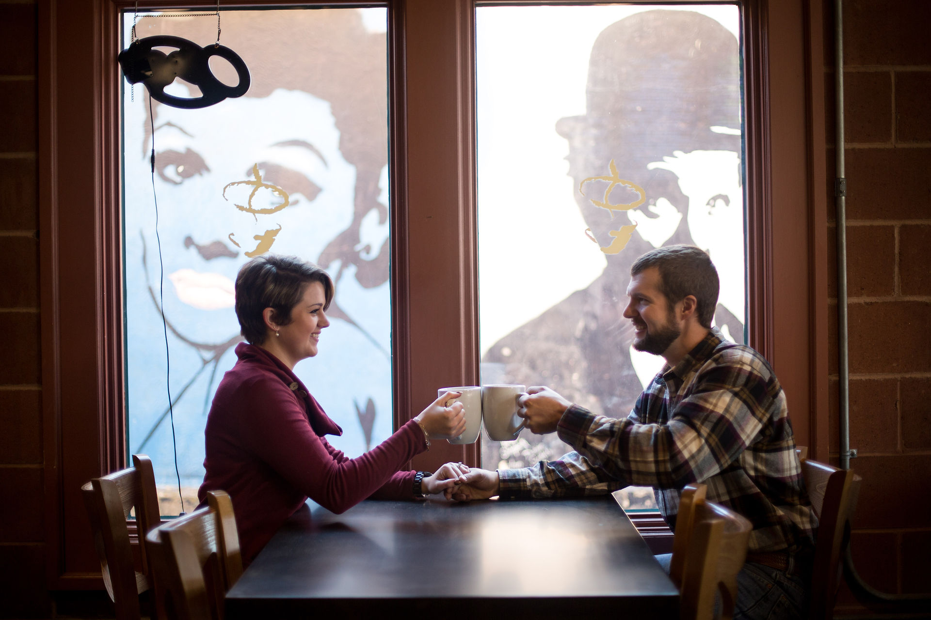 couple sits at a table across from each other cheers-ing coffee cups with murals of charlie chaplin and audrey hepburn visible through the window behind them