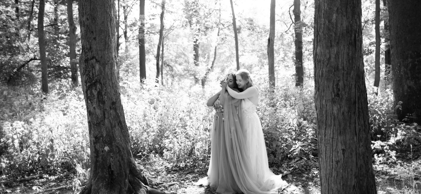 two brides in the woods standing back to front and kissing hands