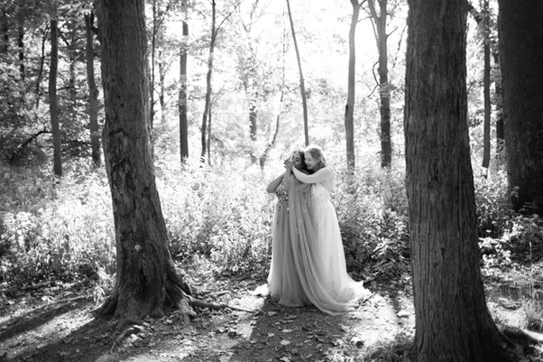 black and white photo of two brides standing in a field with one behind the other kissing hands