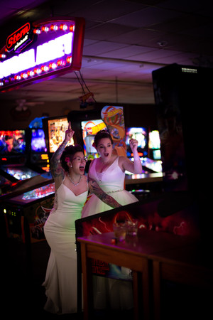 lesbian brides standing at pinball machine wearing wedding dresses and cheering with their arms in the air